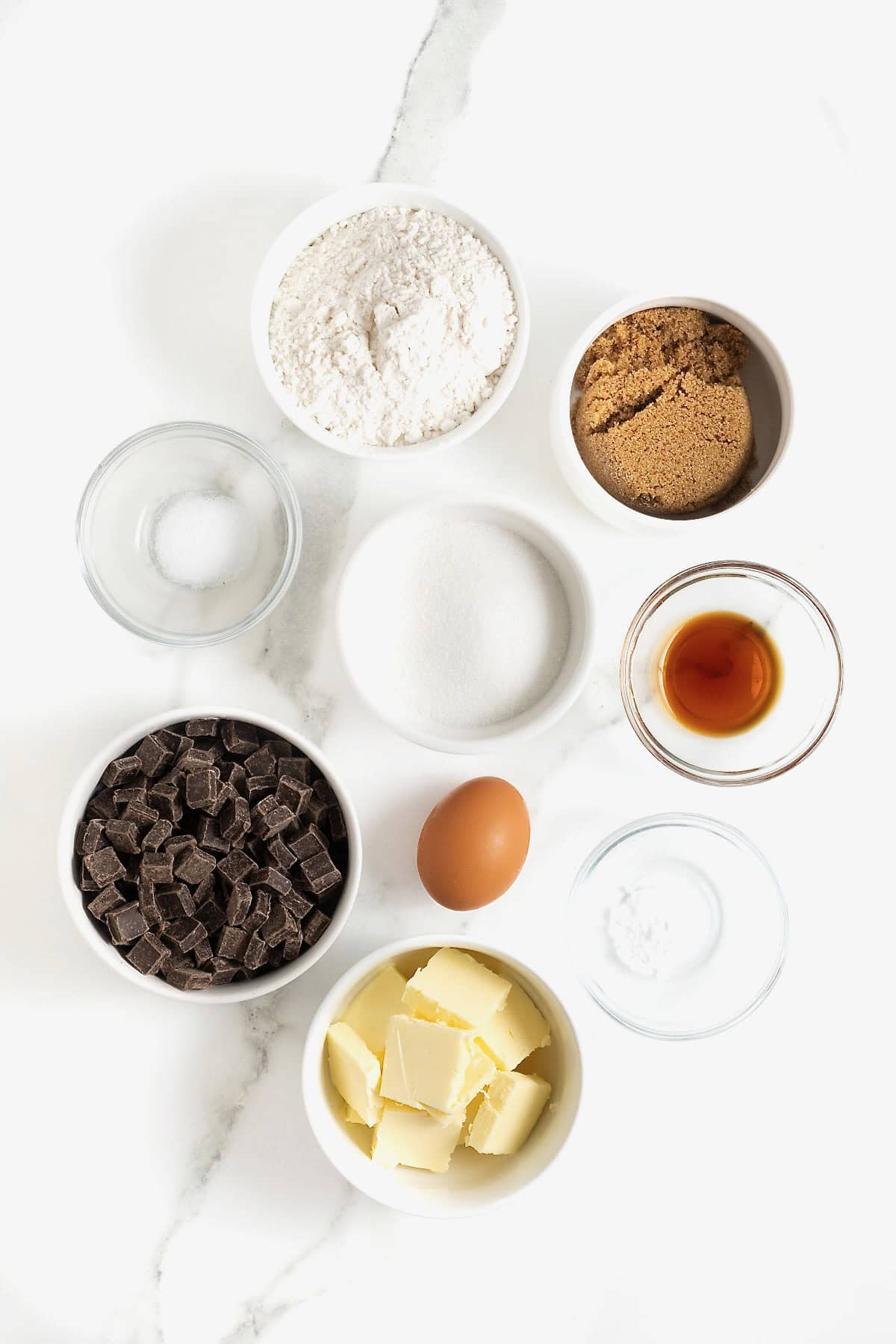 Ingredients to make a skillet chocolate chunk cookie in small glass dishes on a white marble counter.