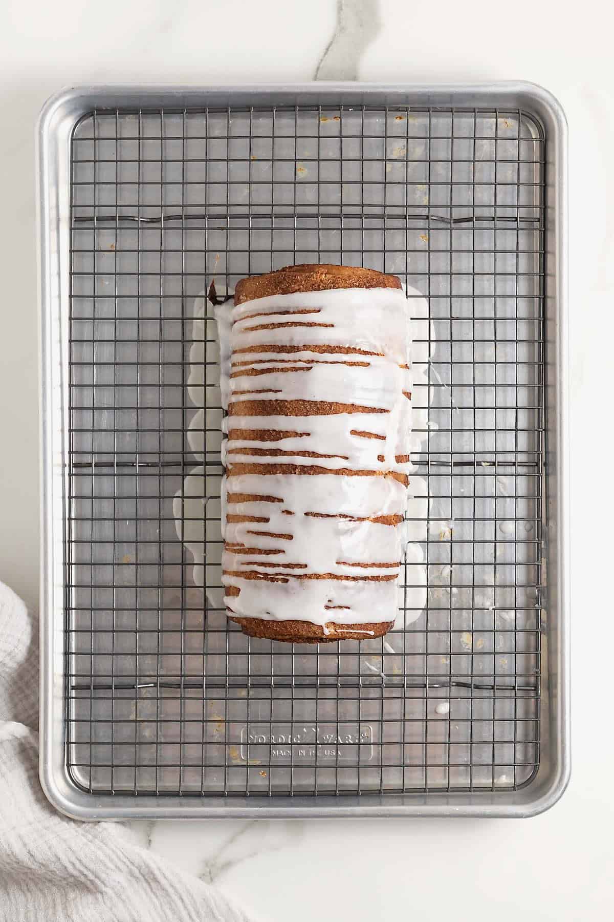 A loaf of cinnamon roll bread with white glazing on a metal cooling rack over an aluminum baking sheet.