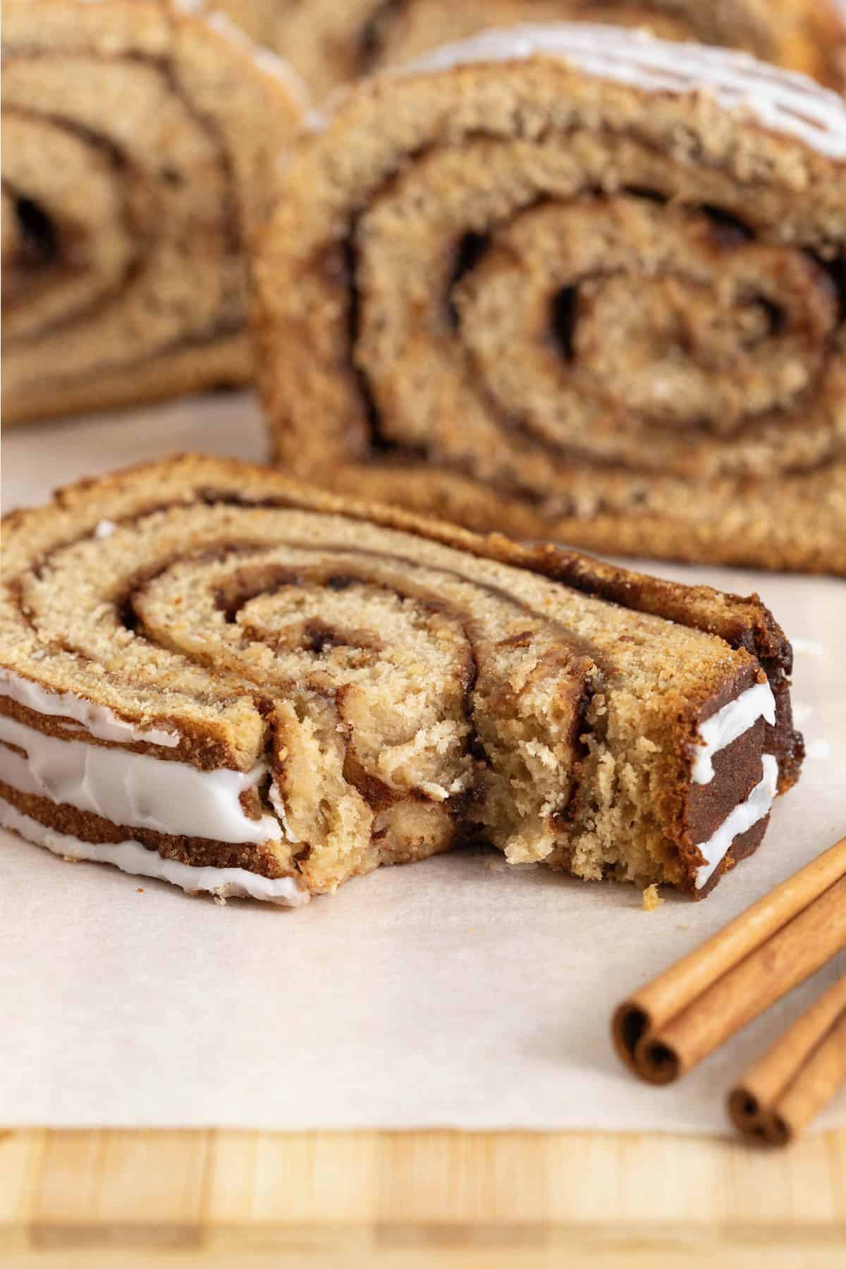 A slice of cinnamon roll bread with a bite taken out of it on a white surface. There are two cinnamon sticks to the bottom right of the photo.