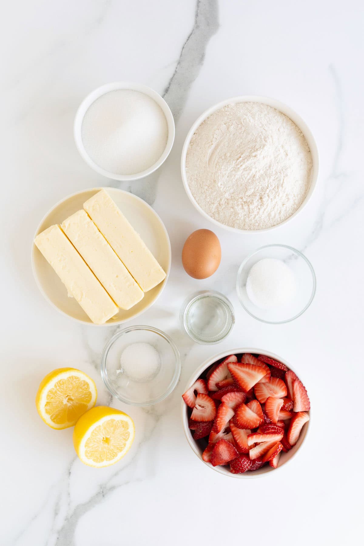 Ingredients to make a strawberry slab pie in white and clear glass dishes on a white marble counter.