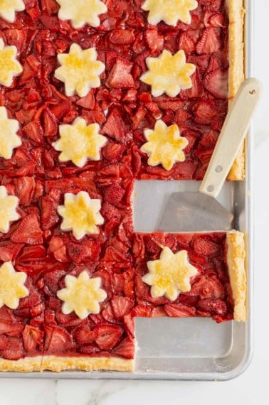 A strawberry slab pie baked in an aluminum rimmed baking sheet. There is a wood handled spatula under one piece of pie.