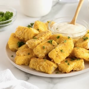 A large white serving plate with a pile of golden battered fish sticks surrounding a small glass dish of tartar sauce.