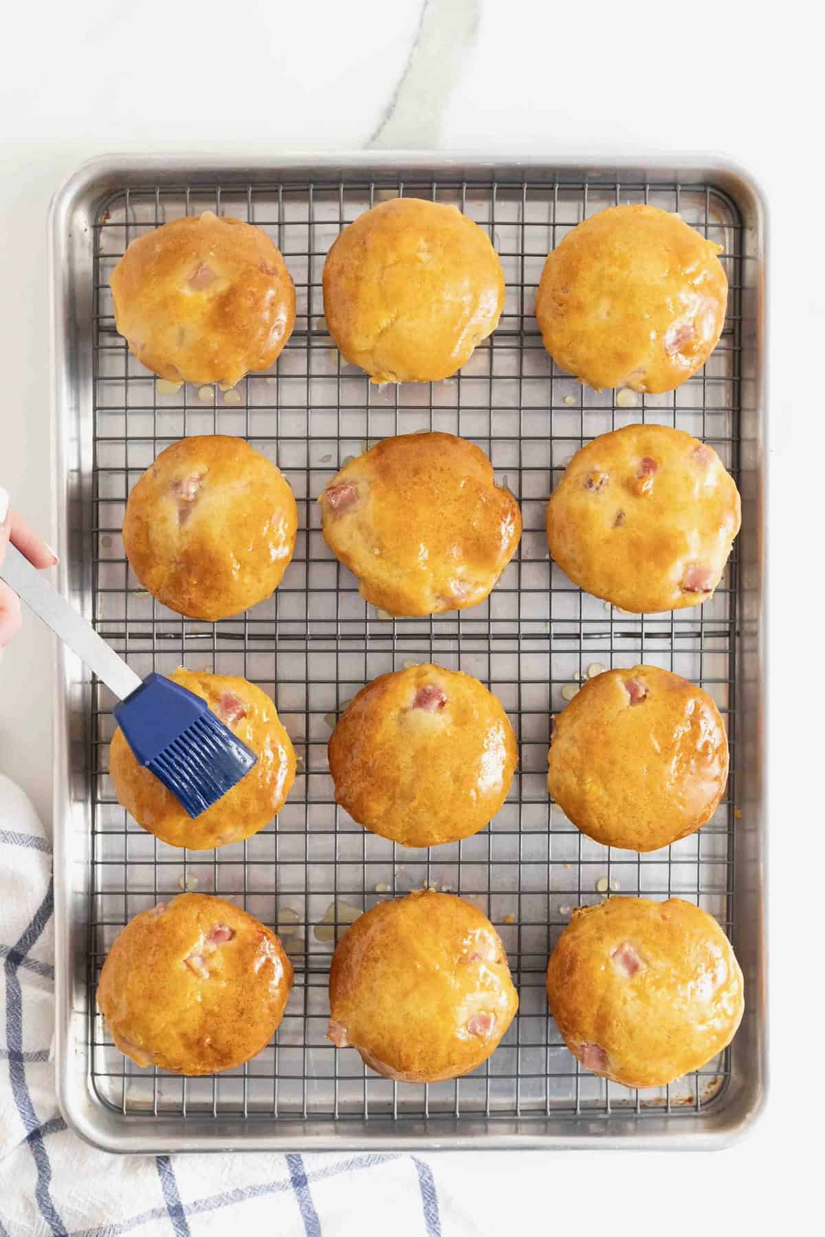 Honey being brushed onto 12 ham and cheddar muffins resting on a wire cooling rack over a baking sheet.