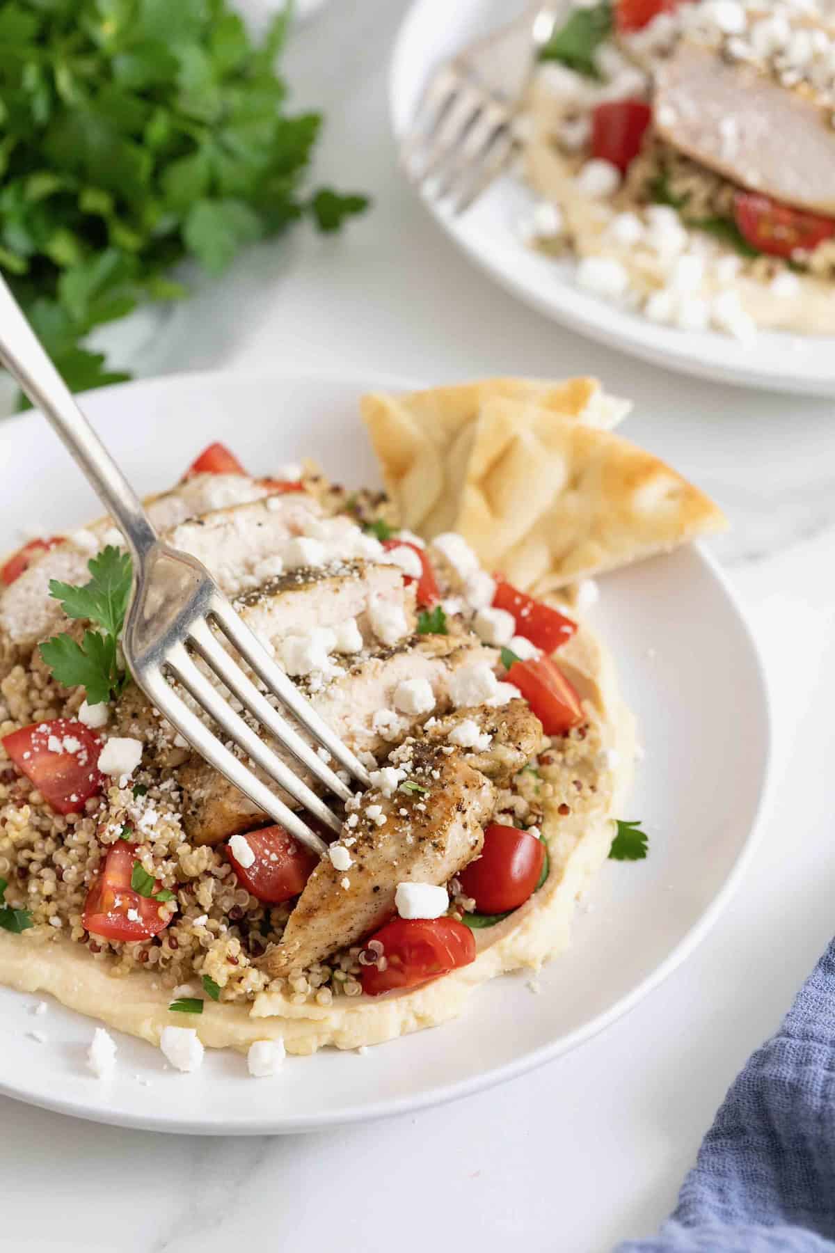A fork piercing a bite of chicken from a hummus quinoa salad on a white salad plate.
