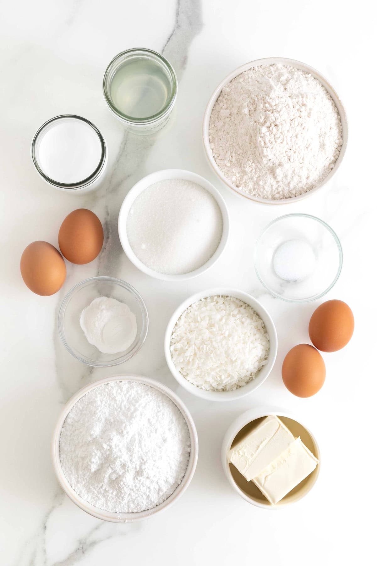 Ingredients to make coconut cream cake in small white and clear glass dishes on a white marble counter.