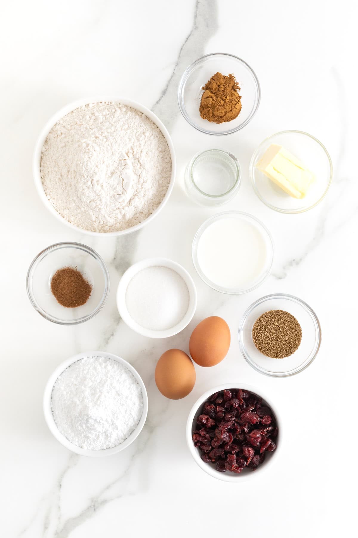 Ingredients to make hot cross buns with dried cranberries in small glass dishes on a white marble counter.