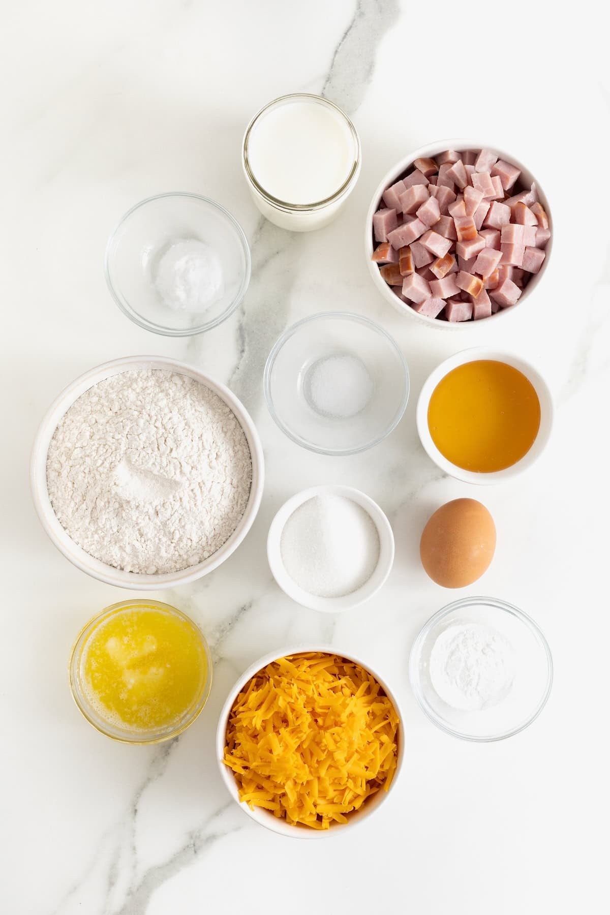 Ingredients to make honey glazed ham and cheddar muffins in small glass dishes on a white marble counter.