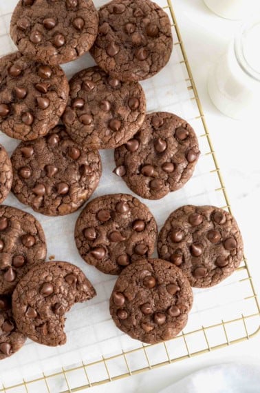 Chocolate cookies with chocolate chips piled on a parchment lined gold-toned cooling rack.