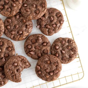 Chocolate cookies with chocolate chips piled on a parchment lined gold-toned cooling rack.