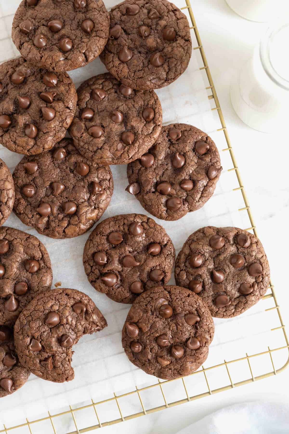 Chocolate cookies with chocolate chips piled on a parchment lined gold-toned cooling rack.