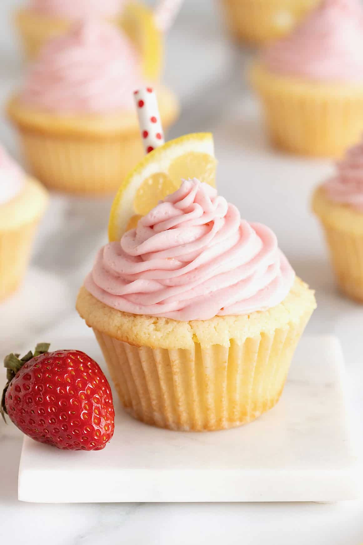 A strawberry lemonade cupcakes on white stone surface. There is a whole strawberry to the left of the cupcake.