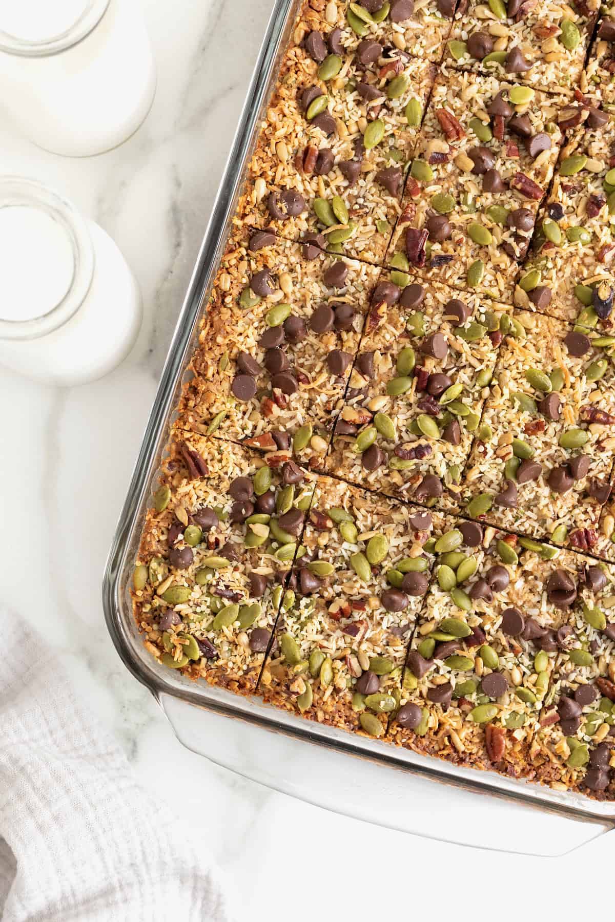 A glass baking dish of granola bars topped with coconut flakes, dark chocolate chips and a mixture of chopped nuts and seeds.