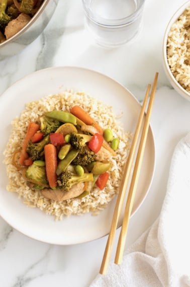A bed of rice on a white plate topped with stir fried vegetables and chicken with a pair of natural wood chopsticks resting on the plate.