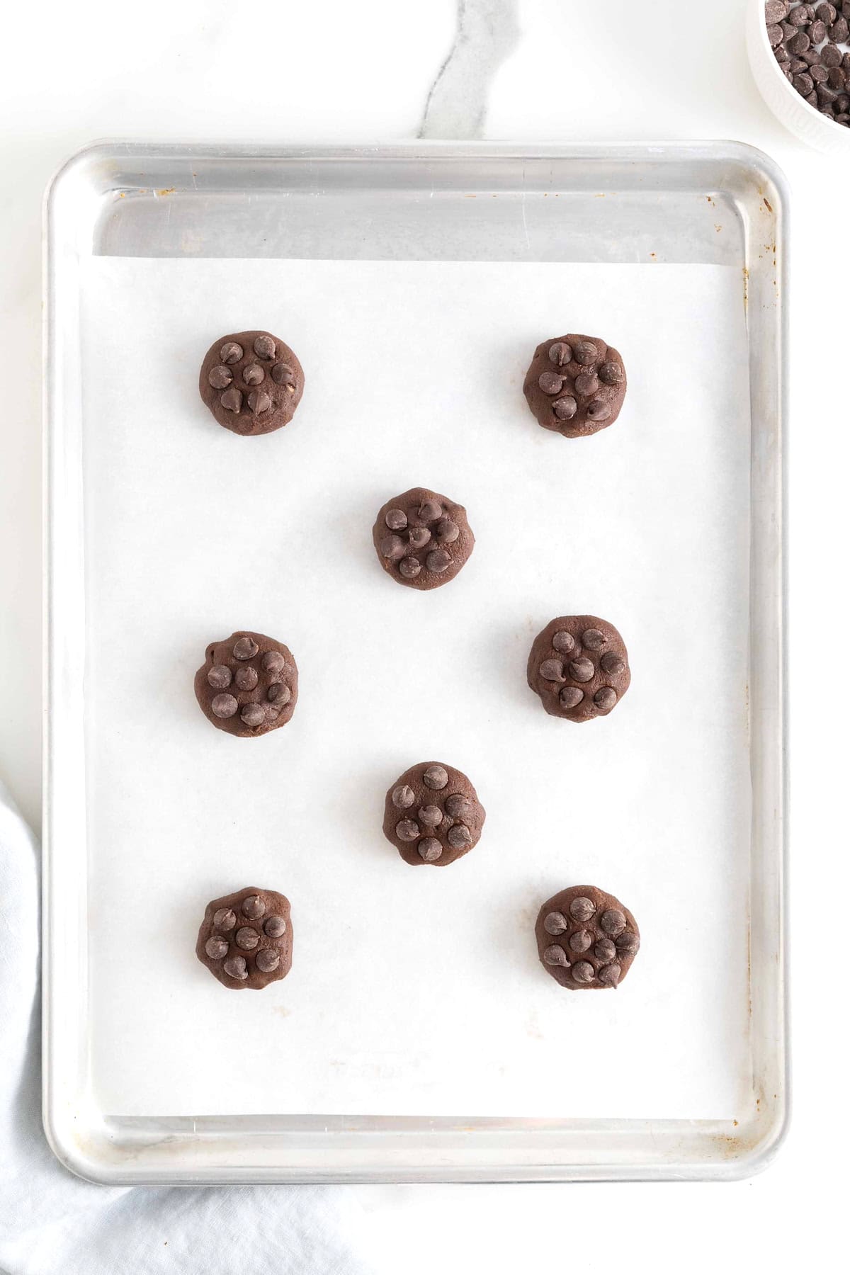 Eight chocolate chocolate chip cookie dough balls on a parchment lined baking sheet.