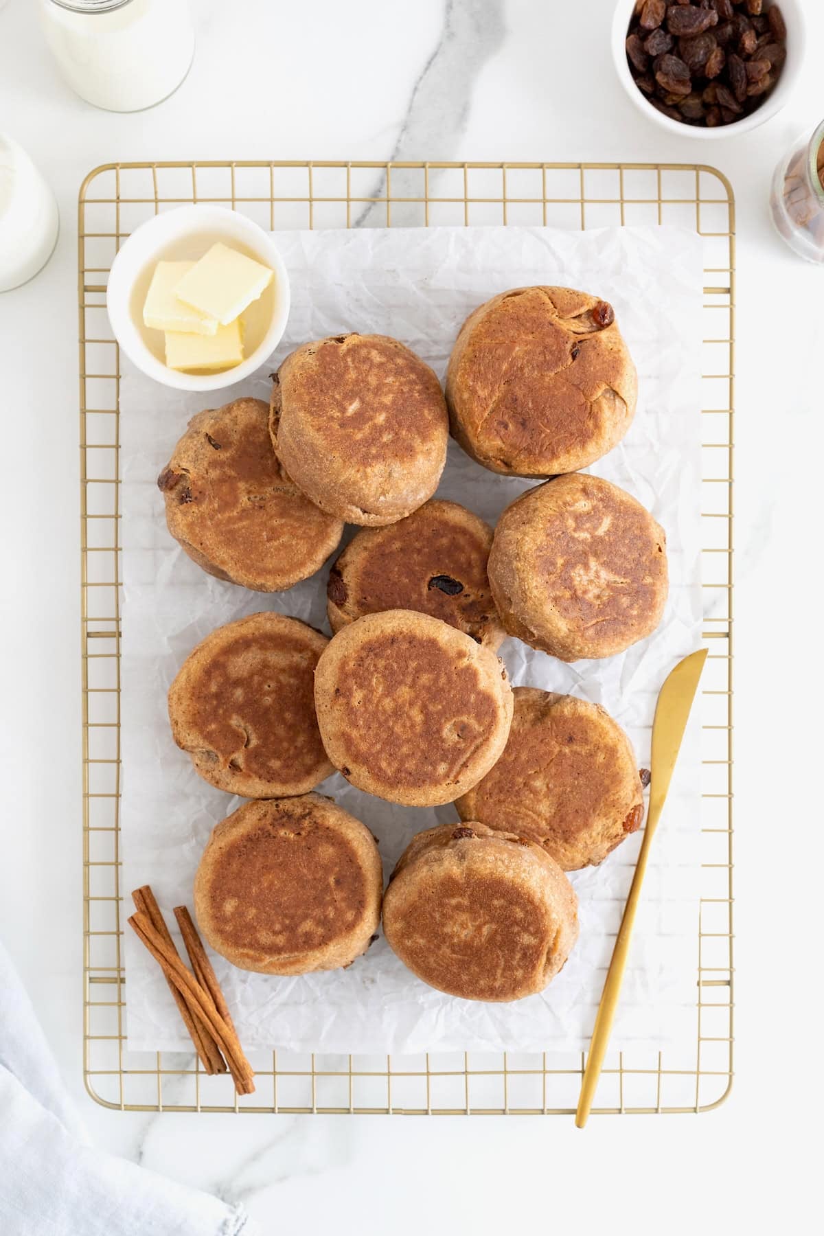 A pile of cinnamon raisin English muffins on a parchment lined gold tone cooling rack.