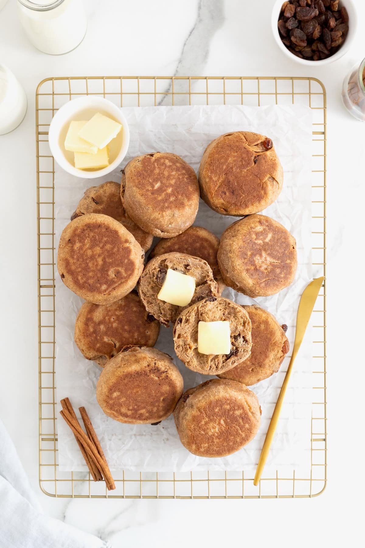 A pile of cinnamon raisin English muffins on a parchment lined gold tone cooling rack. One English muffin is cut in half with butter on it.
