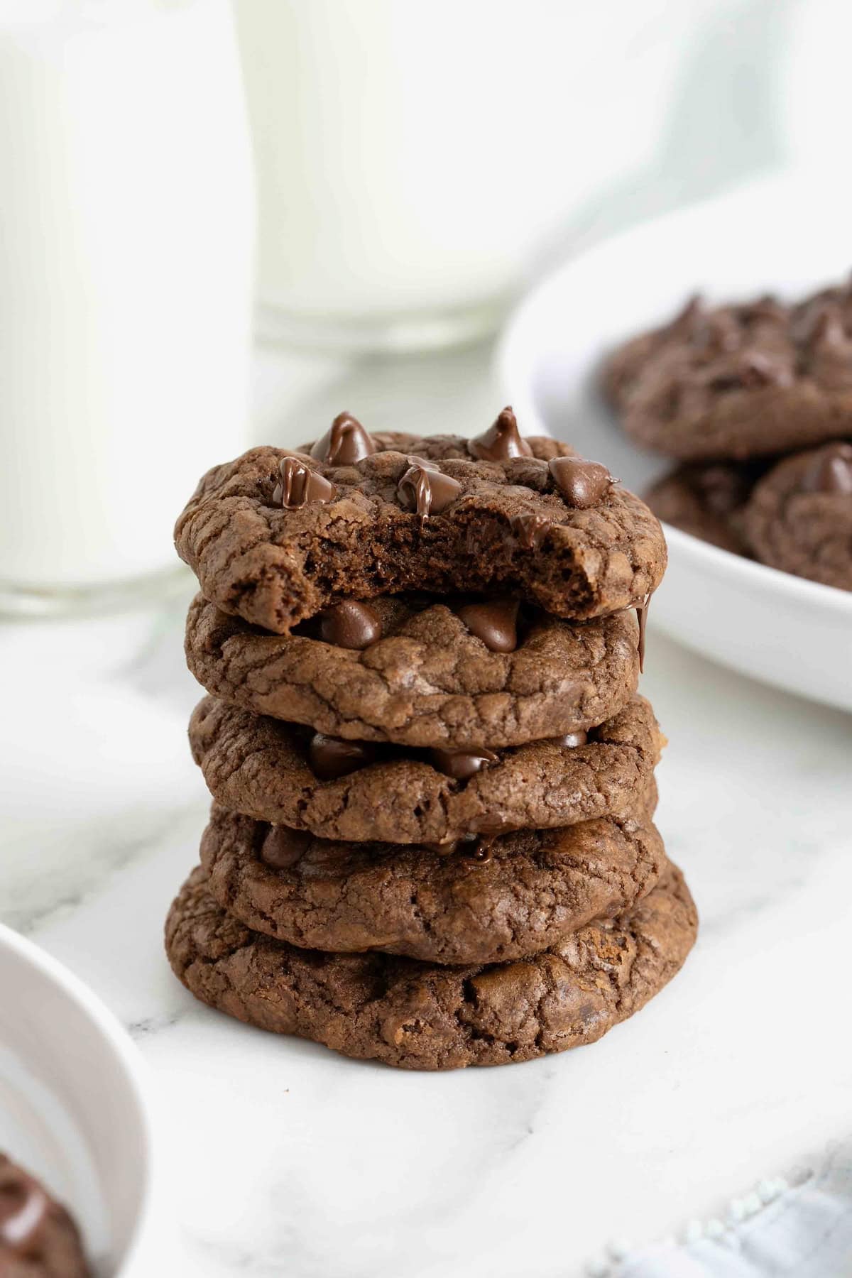 A stack of four double chocolate cookies on a white marble counter. The top cookie has a bit out of it revealing the soft inside.
