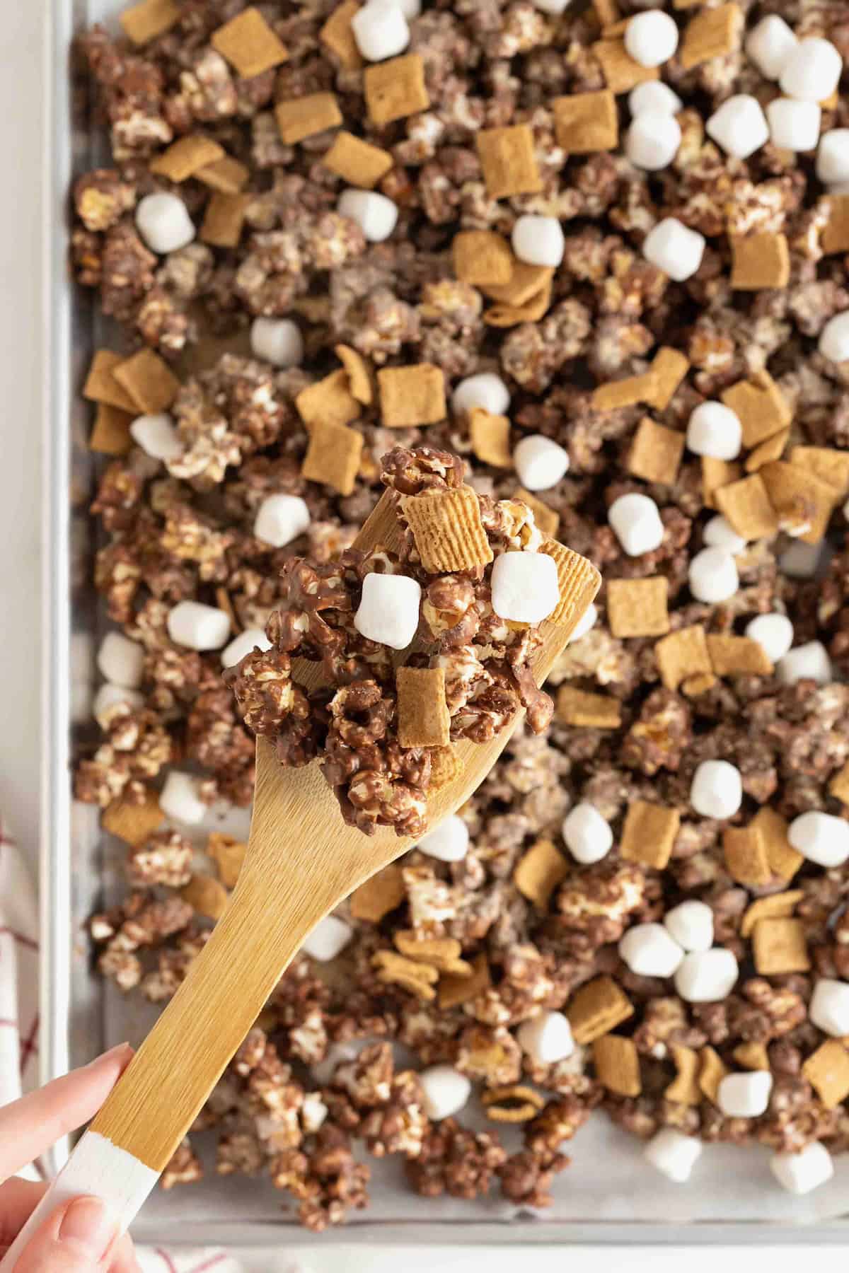 S'mores snack mix on an aluminum baking sheet being scooped up with a wooden spoon.