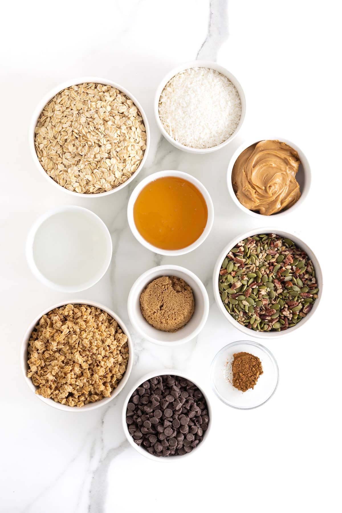 Ingredients to make coconut dark chocolate granola bars in small white dishes on a white marble counter.