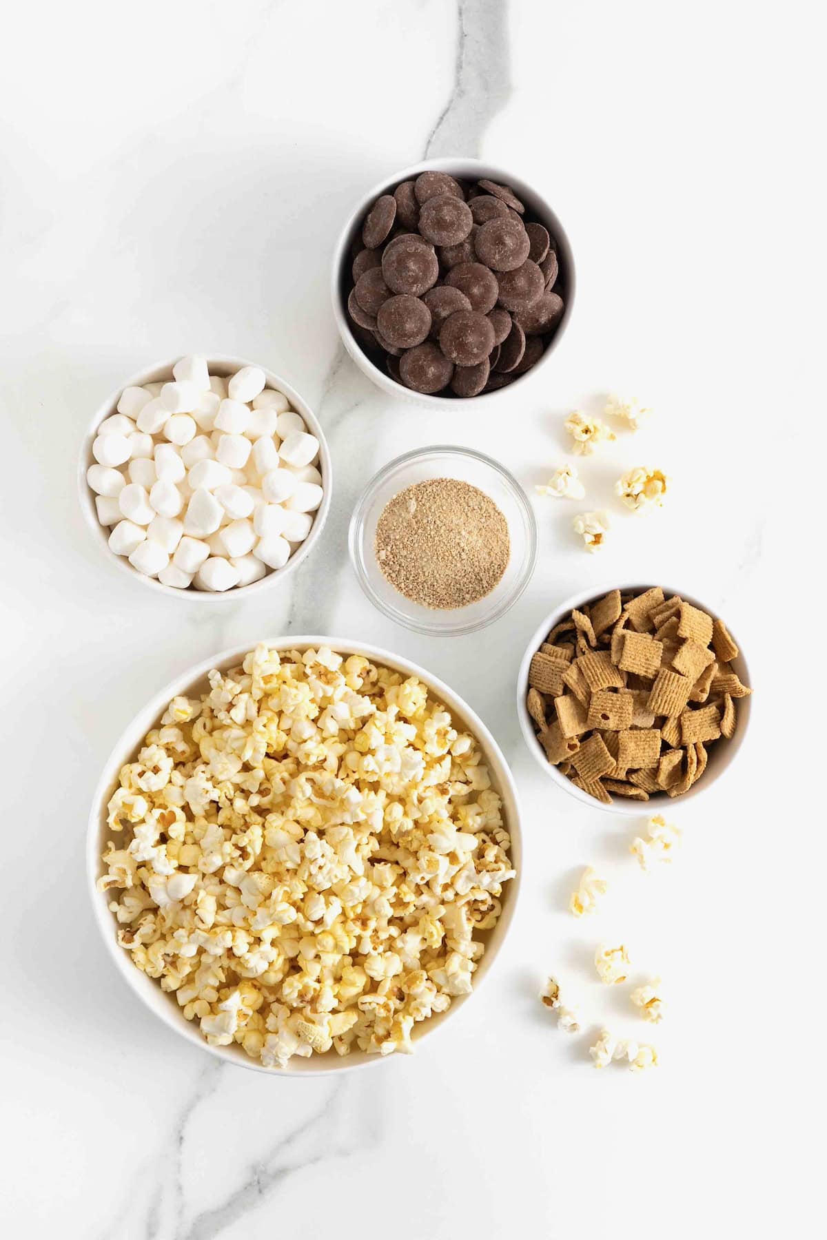Ingredients to make S'mores Popcorn Snack Mix in small white bowls on a white marble counter.
