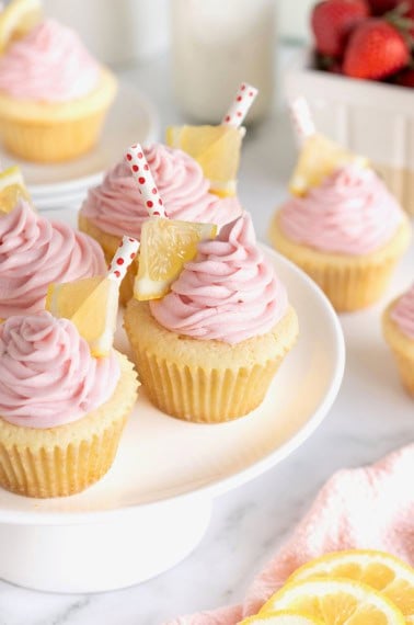 A white cake stand topped with lemon cupcakes with pink frosting garnished with a small triangle of lemon and a red and white polka dot straw.