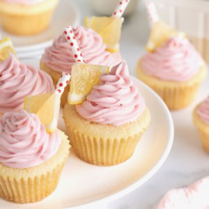 A white cake stand topped with lemon cupcakes with pink frosting garnished with a small triangle of lemon and a red and white polka dot straw.