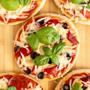 Personal size pizzas topped with shredded mozzarella, sliced black olive and fresh basil on a wooden cutting board.