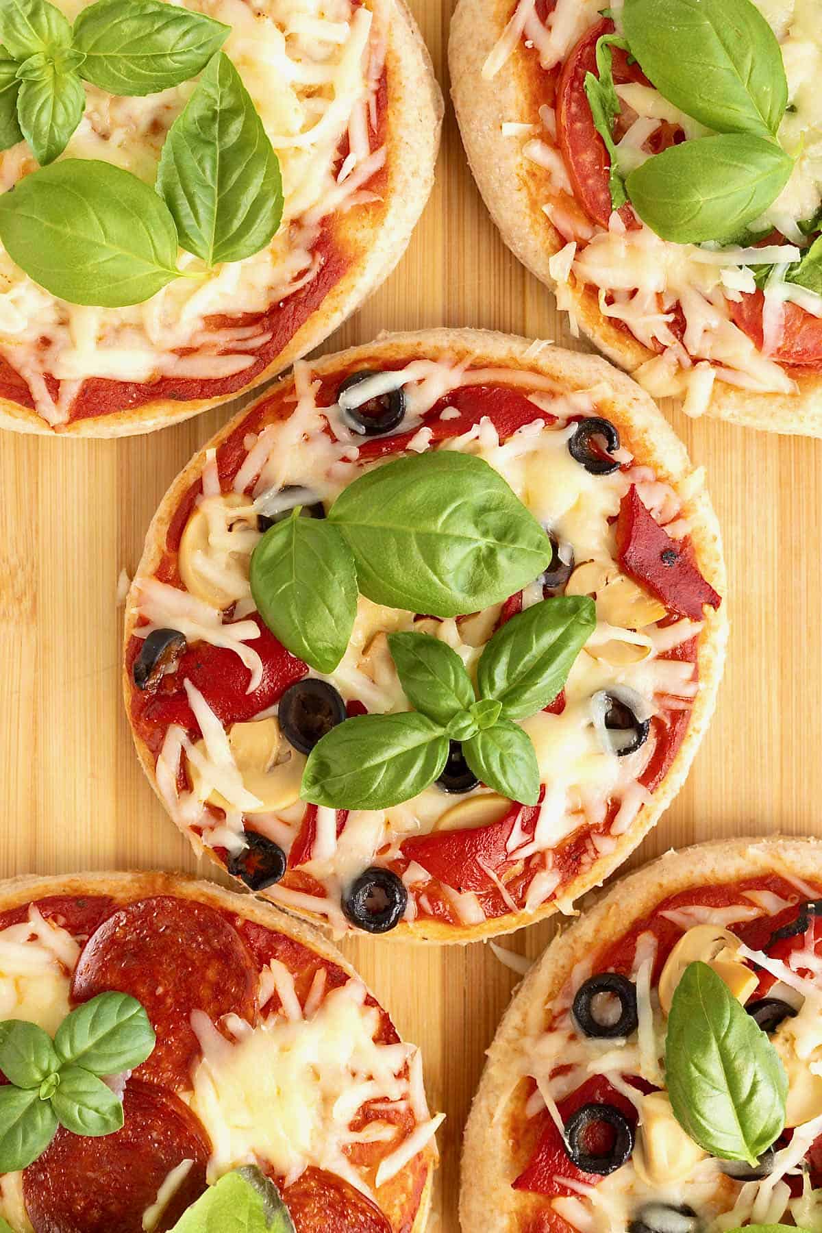 Personal size pizzas topped with shredded mozzarella, sliced black olive and fresh basil on a wooden cutting board.