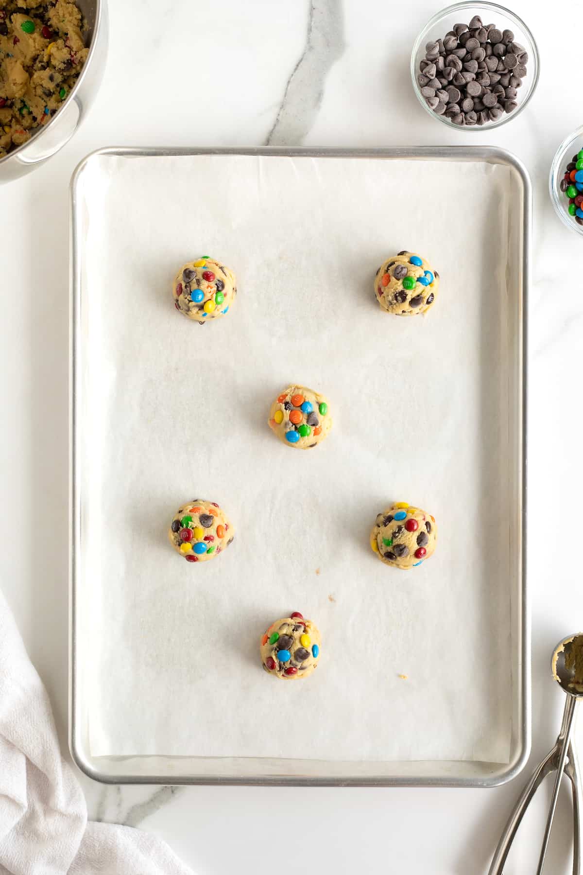 Unbaked balls of M&M chocolate chip cookie dough on a parchment lined baking sheet.