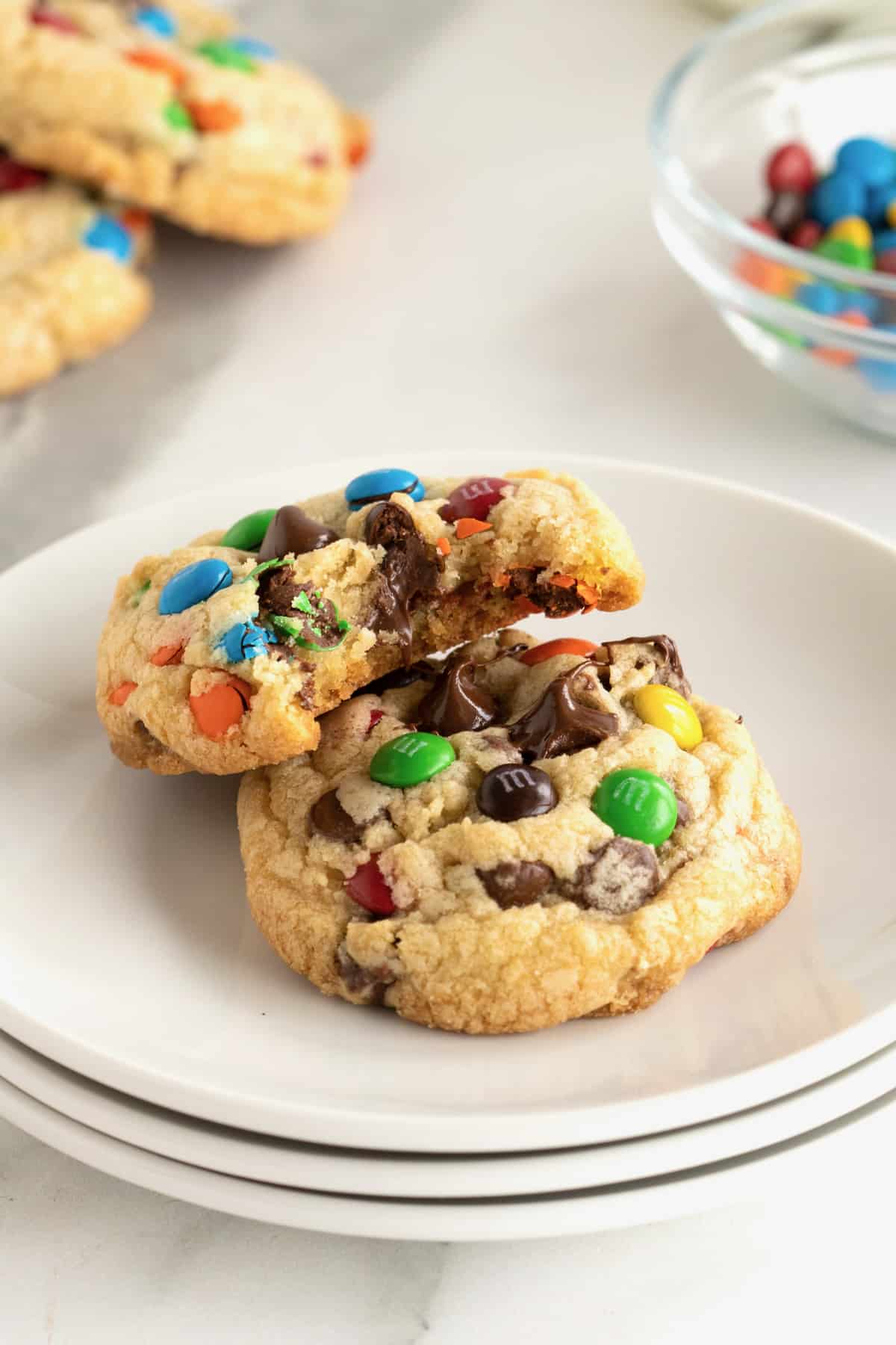 Two chocolate chip M&M cookies on a white plate and one has a bite out of it.
