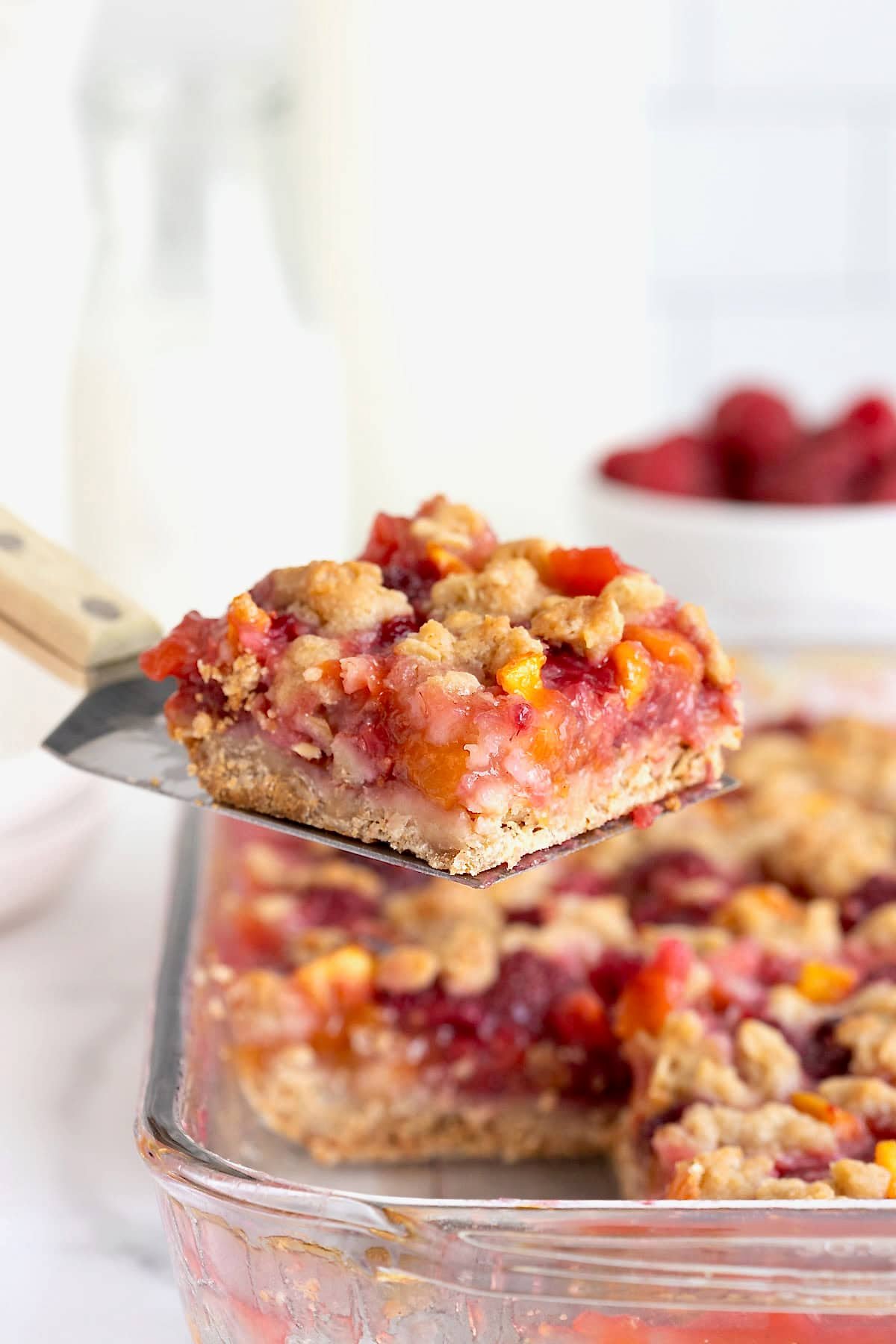 A peach raspberry crumb bar on a spatula being lifted from a baking dish.