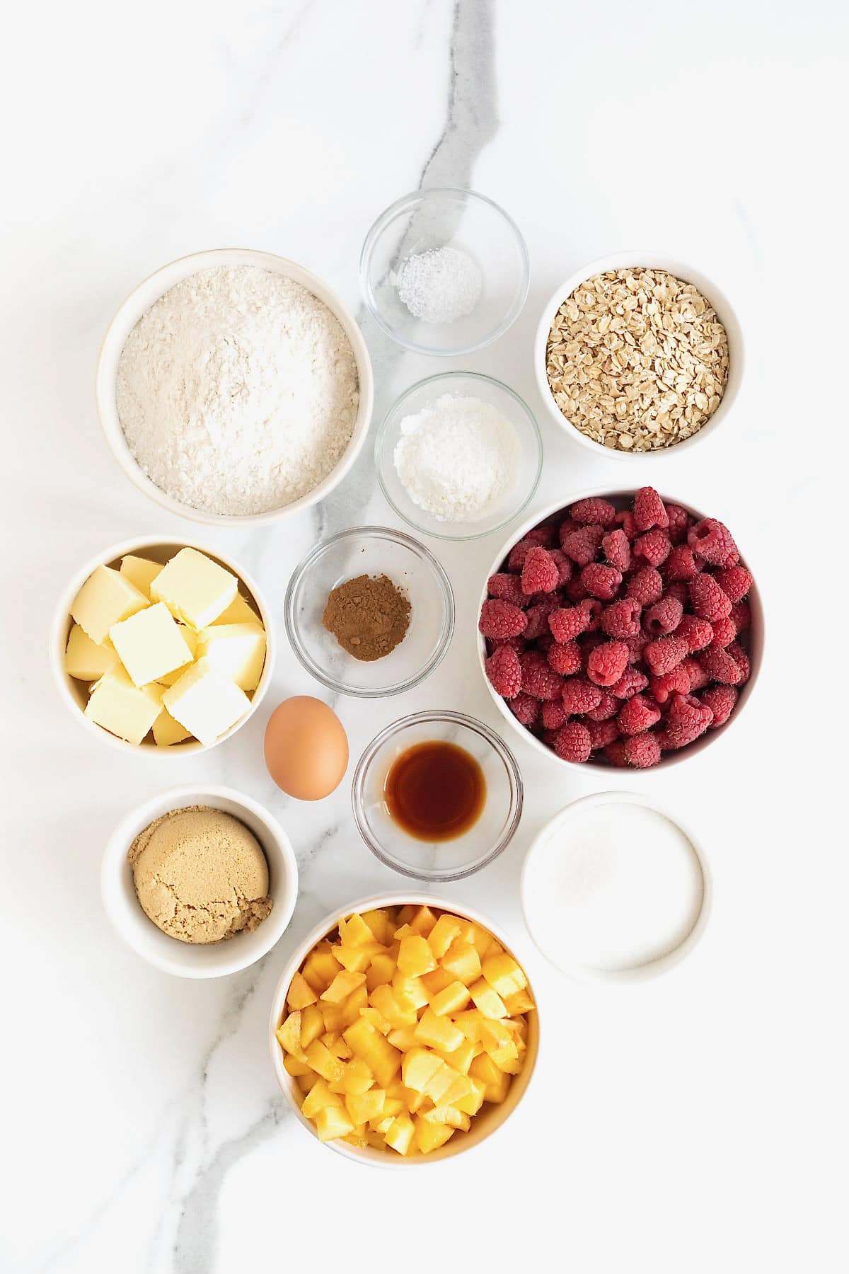 Ingredients to make peach raspberry crumb bars in small glass dishes on a white marble counter.
