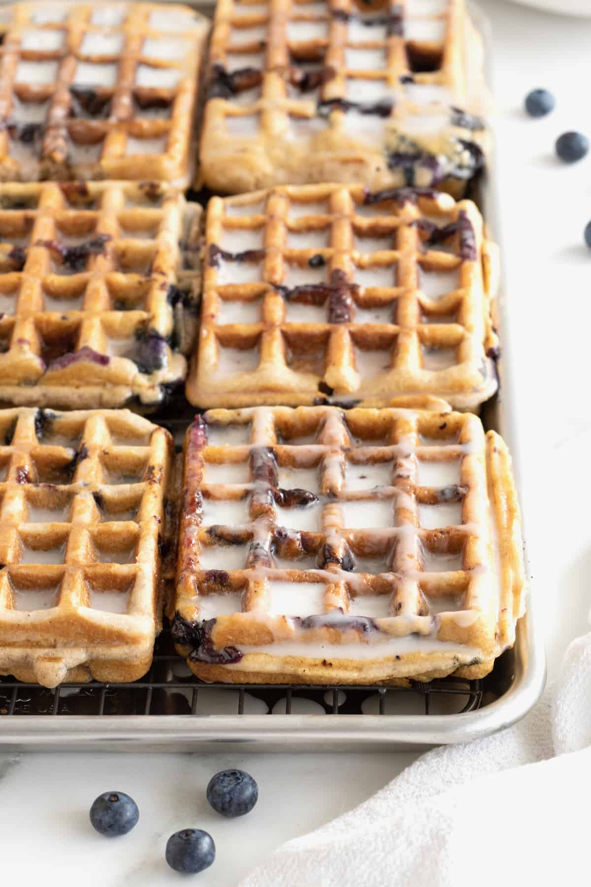 Six blueberry waffles on an aluminum rimmed baking sheet. The waffles have a white glaze on them.