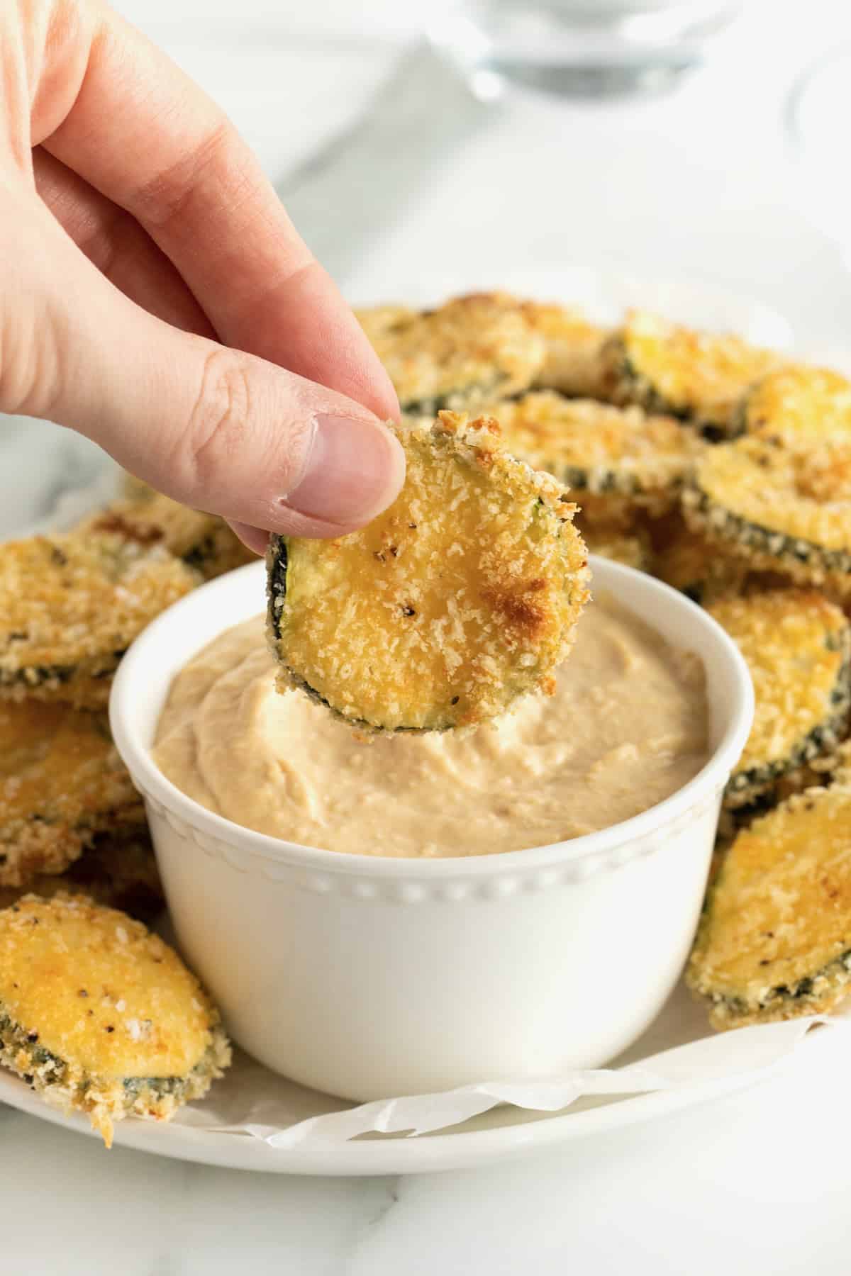 An oblong white serving platter of zucchini chips surrounding a bowl of hummus.