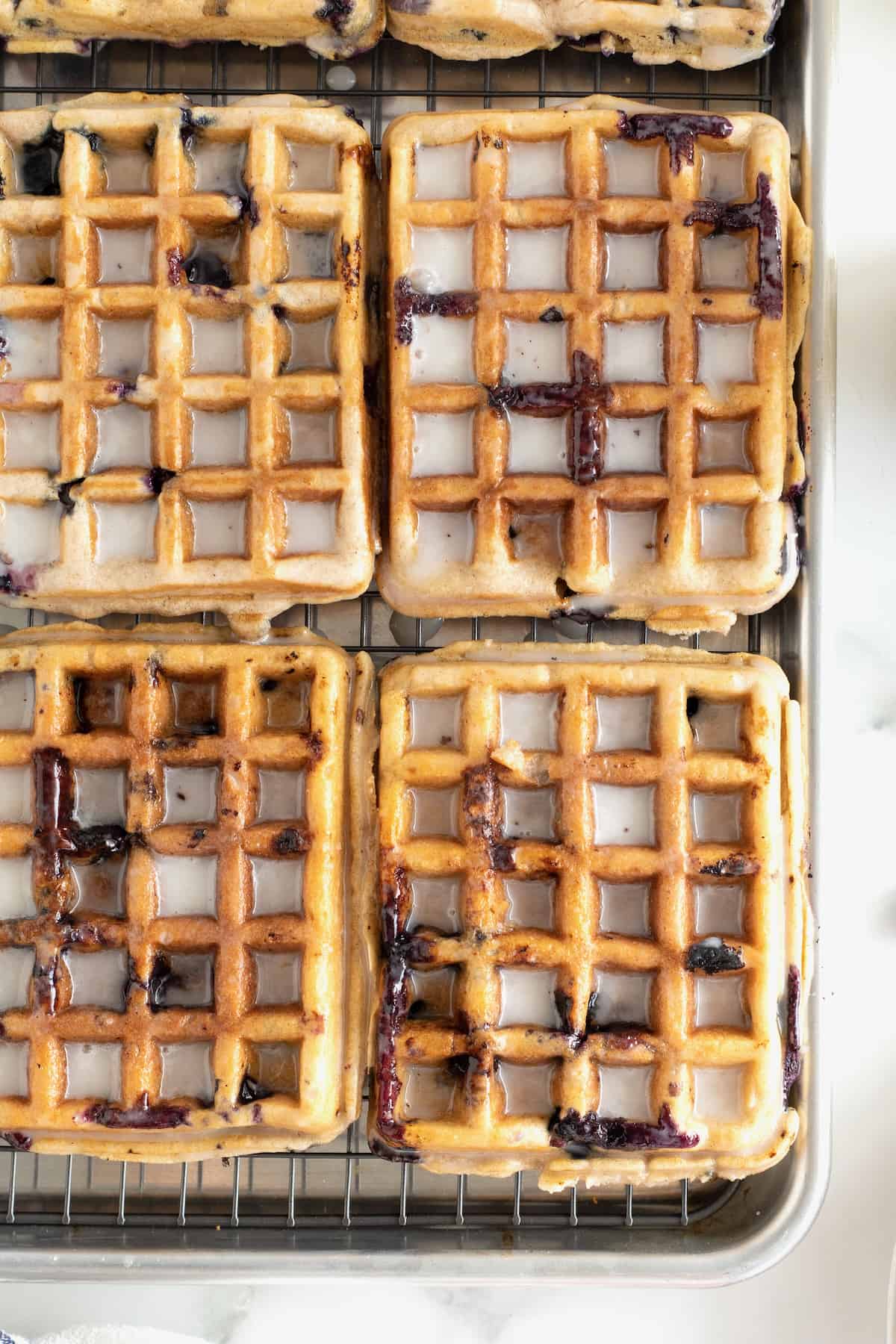Six blueberry waffles on an aluminum rimmed baking sheet. The waffles have a whiteish purple glaze on them.