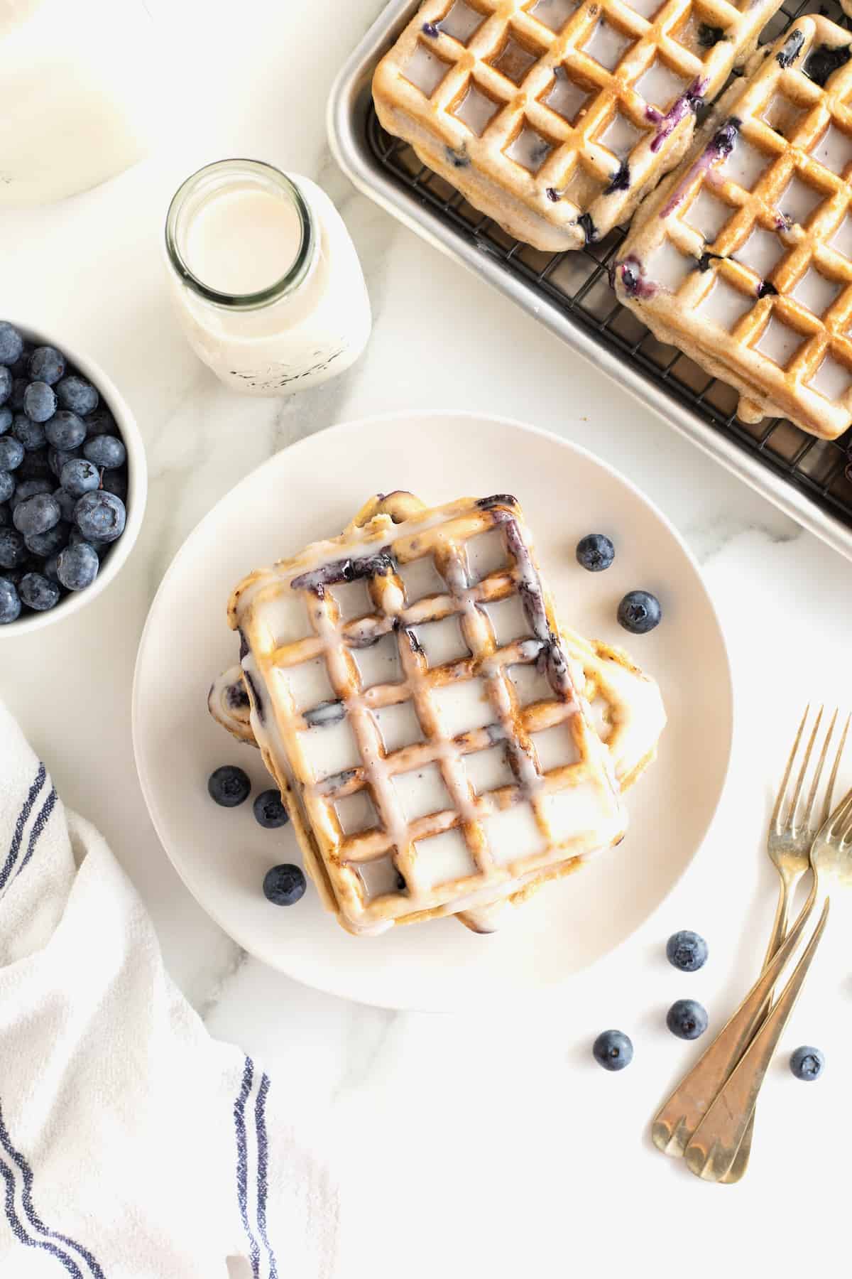 A single blueberry waffle on a white plate with white glaze dripping off and blueberries scattered around it. To the lower right of the plate are two gold toned forks.