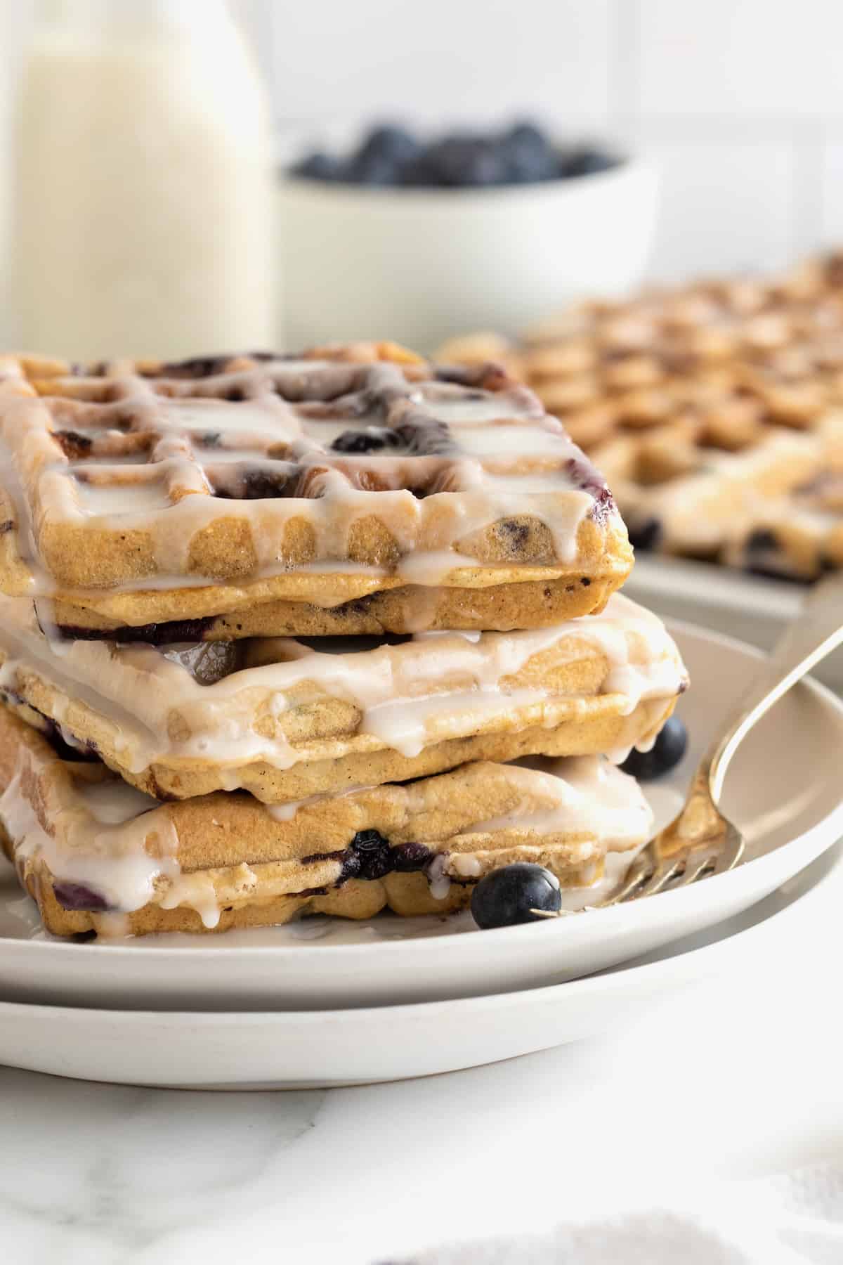 Three glazed blueberry waffles stacked on a small white plate. There is a fork resting on the plate.
