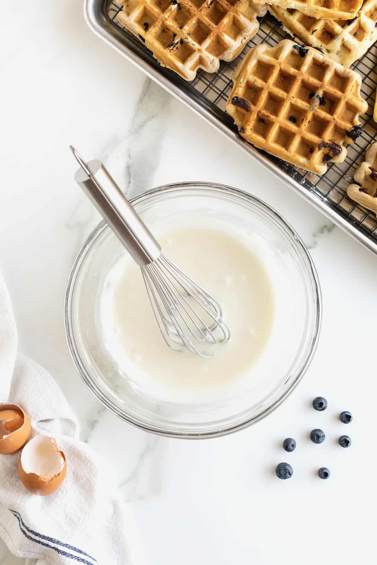 A metal whisk in a glass bowl of white glaze.