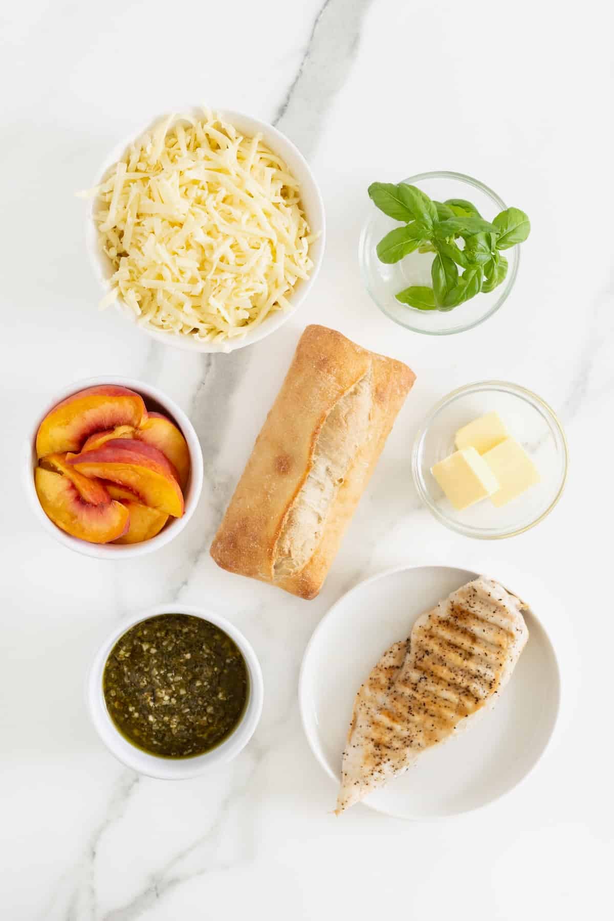 Ingredients to make Chicken Pesto Peach Panini in glass dishes on a white marble counter.