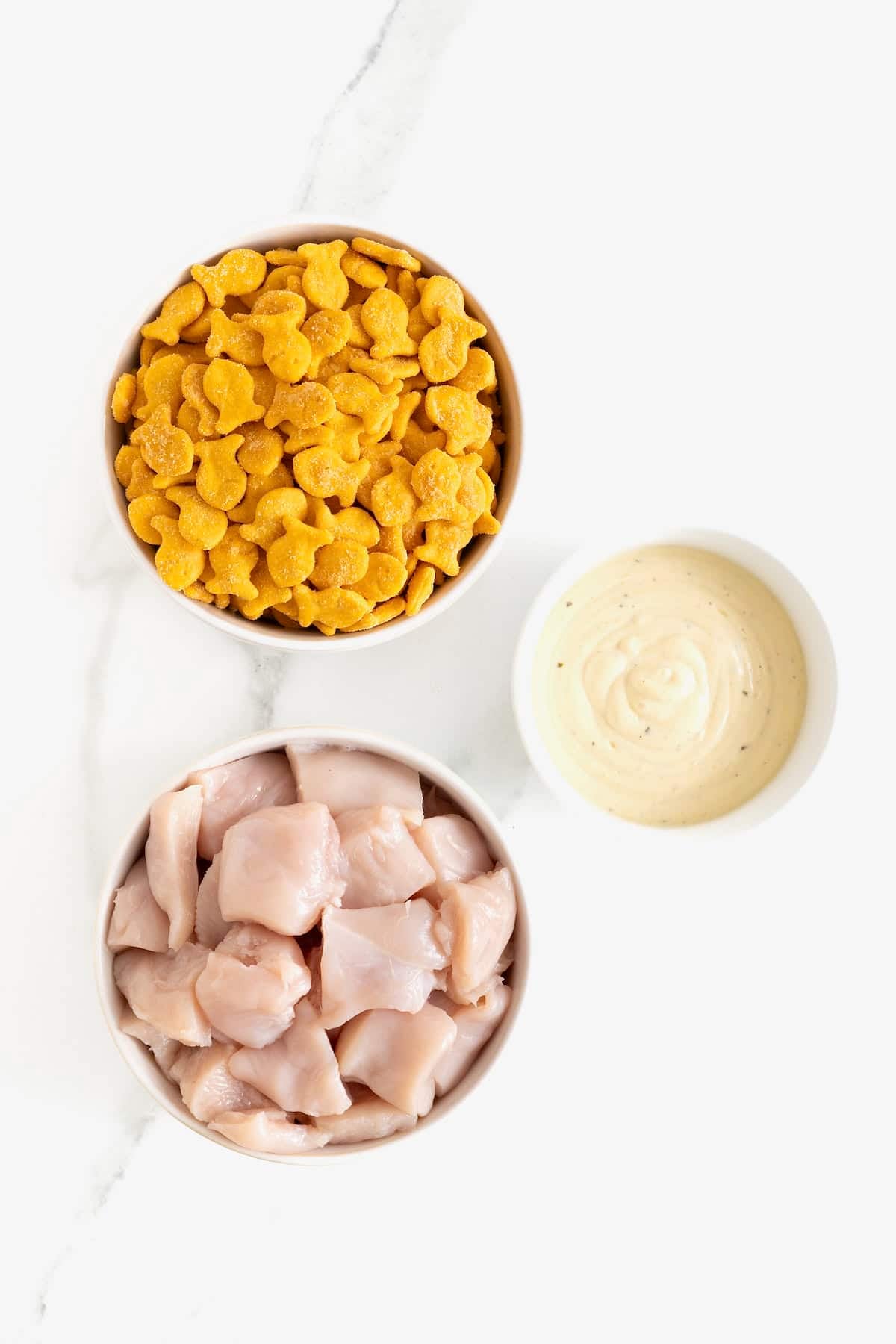 Ingredients to make goldfish chicken nuggets in white dishes on a marble counter.