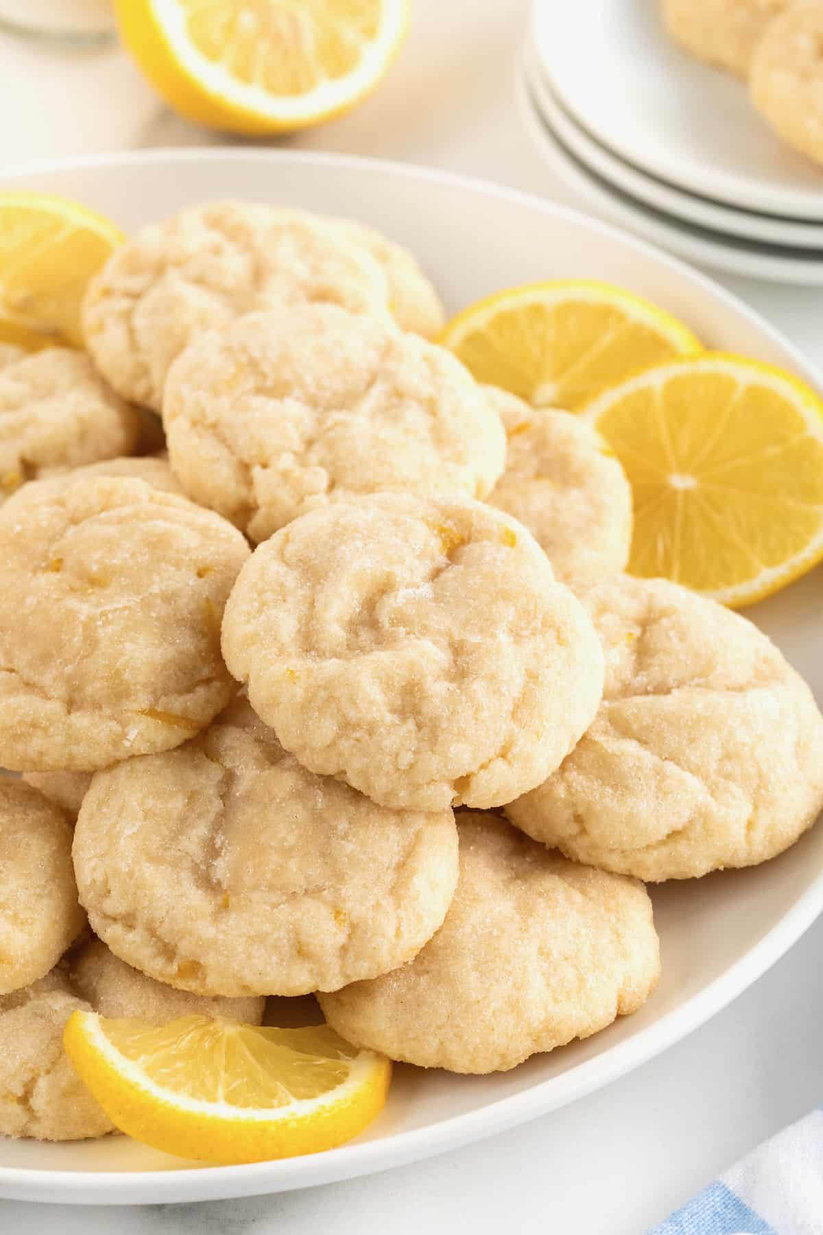 A large white round serving plate piled with lemon cookies dusted with sugar. There are two lemon slices garnishing the plate on the upper right hand.