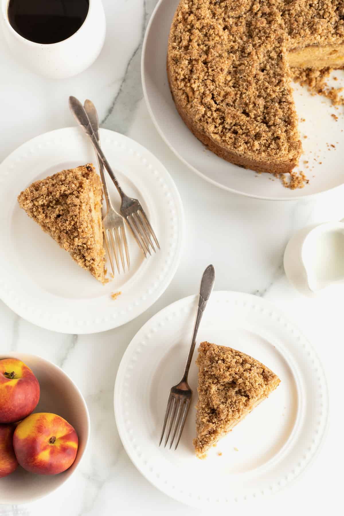 Two slices of peach coffee cake on white plates with forks and fresh peaches in the foreground.