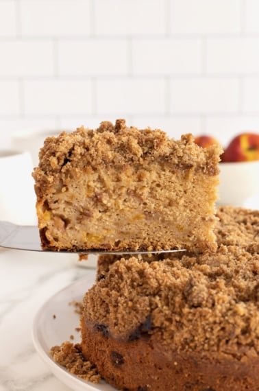 A slice of coffee cake with cinnamon sugar streusel on a pie server with a peach in the background.