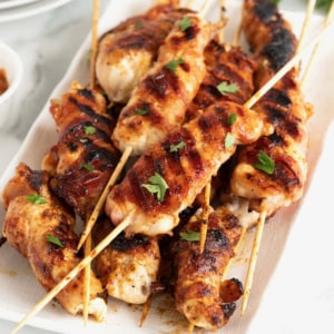 A rectangular white serving platter of grilled chicken on bamboo skewers.