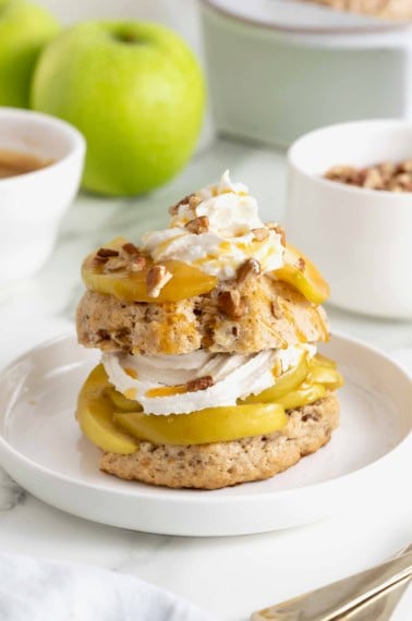 A caramel apple pecan shortcake topped with whipped cream and garnished with pecan pieces on a white rimmed plate.