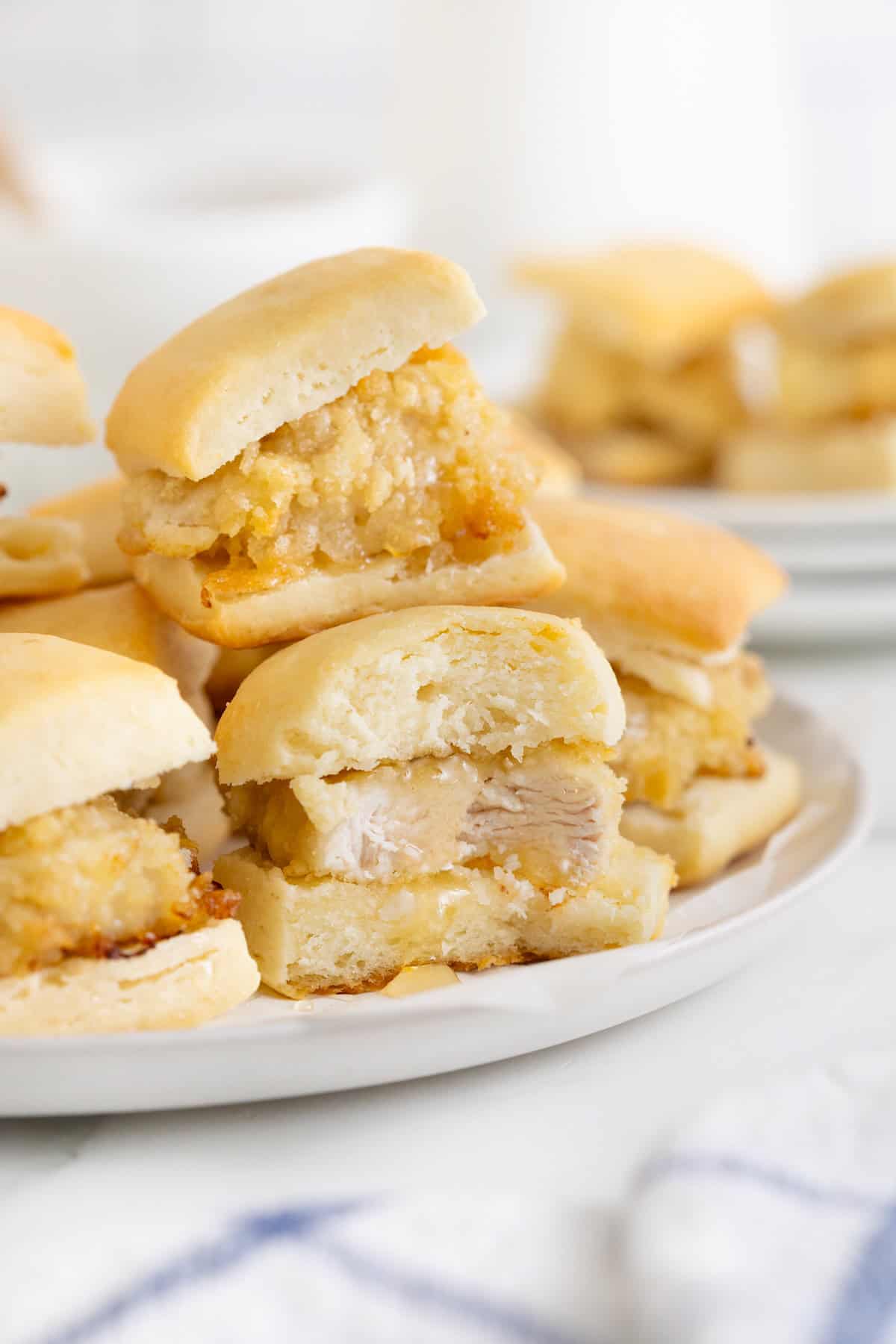 A pile of mini chicken biscuits that are drizzled with honey. The foremost sandwich has a bite out of it.