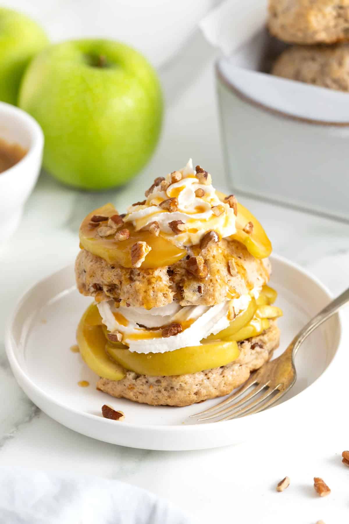A caramel apple pecan shortcake topped with whipped cream and garnished with pecan pieces on a white rimmed plate.