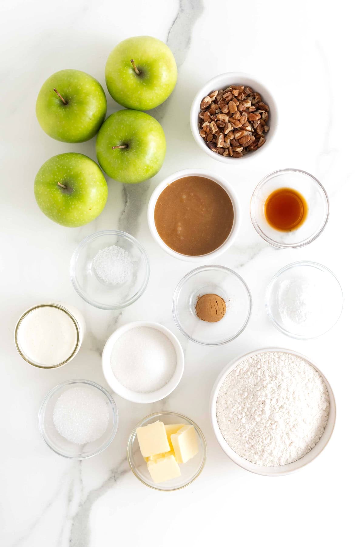 Ingredients to make caramel apple pecan shortcakes in small glass dishes on a white marble counter.
