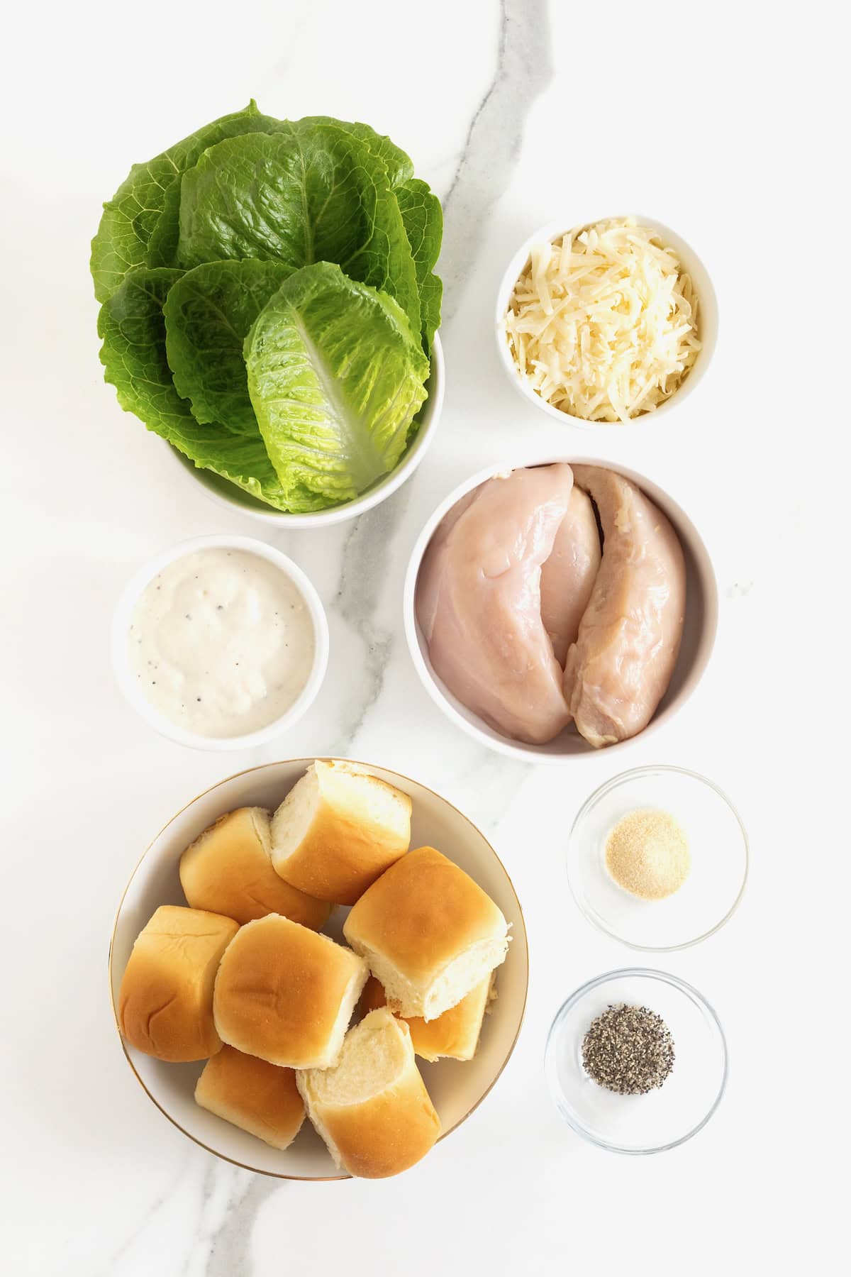 Ingredients to make grilled chicken Caesar sliders on white and clear glass dishes on a white marble counter.