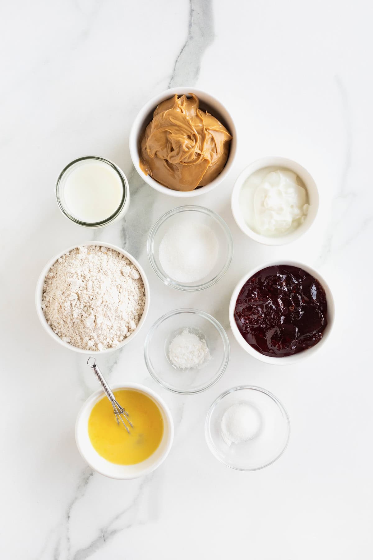 Ingredients to make peanut butter and jelly muffins in small glass dishes on a white marble counter.